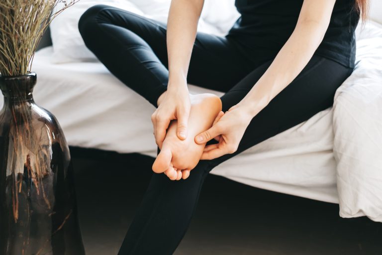 A woman massaging her foot while sitting on a bed, wearing black leggings and a fitted top.